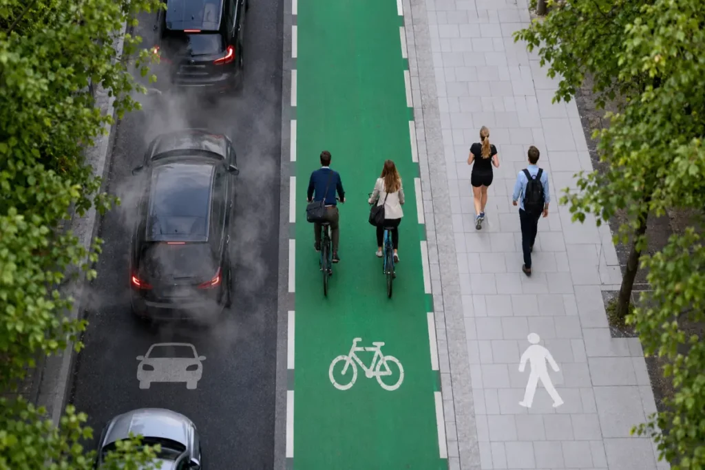 Top-down aerial photograph showing active transportation reducing carbon emissions, with a polluted car traffic lane beside a clean green bike lane and pedestrian walkway in a modern European-style city.